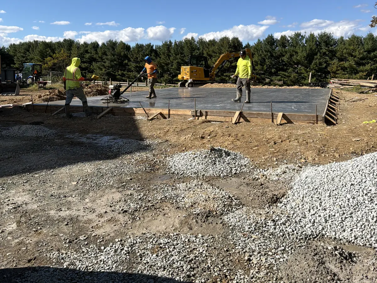 Crew members and a yellow excavator beside a slab in progress, with gravel in the foreground