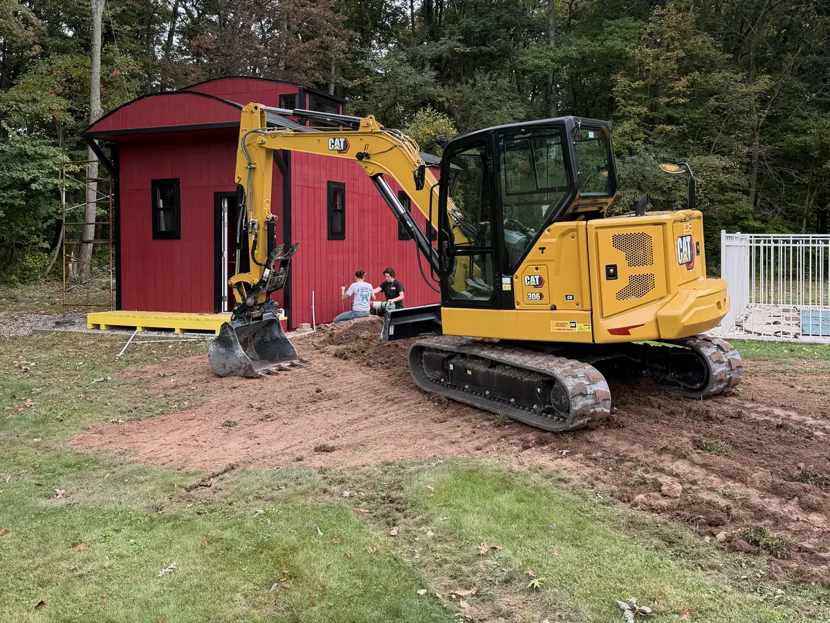 Yellow excavator on disturbed ground beside a red outbuilding